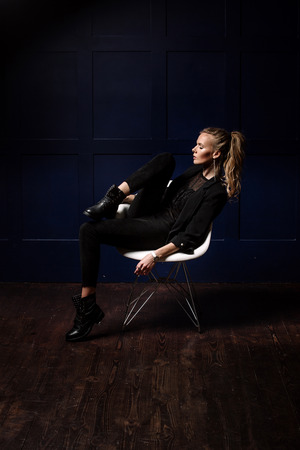 Sexy young model in dark clothes, sitting on a white chair and posing on a dark blue background. Watch on hand. Fashion, Beauty, Styleの写真素材