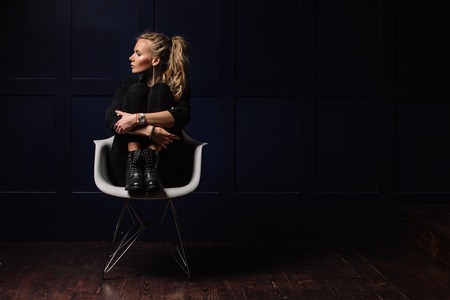 Sexy young model in dark clothes, sitting on a white chair and posing on a dark blue background. Watch on hand. Fashion, Beauty, Styleの写真素材