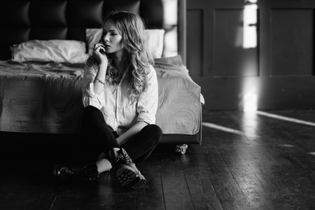 A girl with long blond hair dressed in black jeans, a striped shirt and black shoes sitting on the bed with blue linens. Fashionable casual outfit.の写真素材