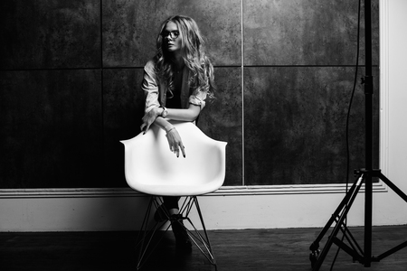 A young beautiful stylish woman in a white shirt, black jeans, glasses in an involuntary pose sits on a white chair on a gray background. Fashion studio style full length portrait of a girl sitting on a chairの写真素材