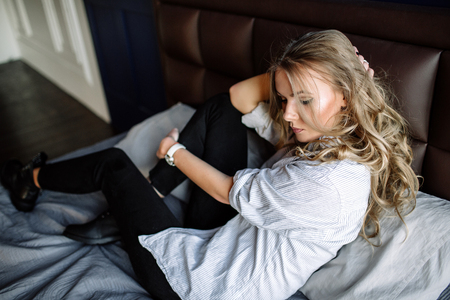 A girl with long blond hair dressed in black jeans, a striped shirt and black shoes sitting on the bed with blue linens. Fashionable casual outfit.の写真素材