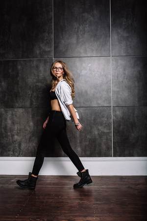 Fashion style of fashion. Fashionable woman in stylish clothes posing on a gray background in the studio. A beautiful sexy girl wearing black glasses, black jeans, black shoes, a white striped shirt.の写真素材