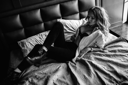 A girl with long blond hair dressed in black jeans, a striped shirt and black shoes sitting on the bed with blue linens. Fashionable casual outfit.の写真素材