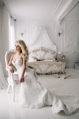Wedding. Bride in a beautiful dress indoors in a white studio, like at home. The fashionable wedding style is taken in full length. Young attractive blond model with curly hair, like a brideの写真素材
