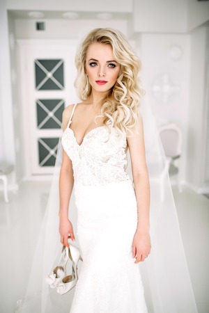Wedding. Bride in a beautiful dress indoors in a white studio, like at home. The fashionable wedding style is taken in full length. Young attractive blond model with curly hair, like a brideの写真素材