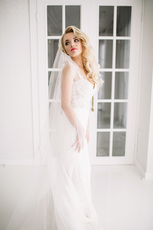 Wedding. Bride in a beautiful dress indoors in a white studio, like at home. The fashionable wedding style is taken in full length. Young attractive blond model with curly hair, like a brideの写真素材