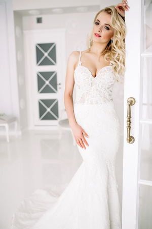 Wedding. Bride in a beautiful dress indoors in a white studio, like at home. The fashionable wedding style is taken in full length. Young attractive blond model with curly hair, like a brideの写真素材