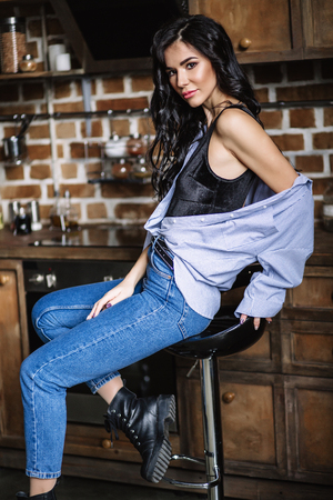 Beautiful brunette in the kitchen, modern housewife, fashion, wooden brown furniture, dressed in blue jeans and a blue shirtの写真素材