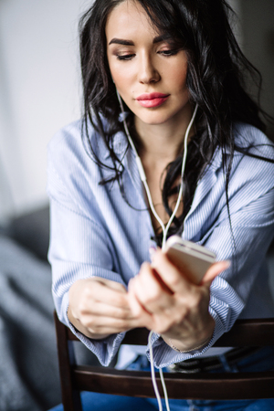 A beautiful brunette listens to music in headphones in a room near the sofa, is dressed in a shirt and blue jeans, blueの写真素材