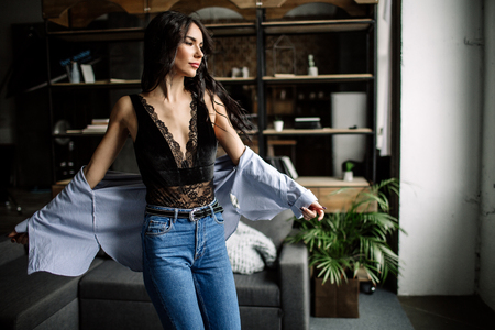 Foreground. Beautiful brunette dressed in a blue shirt and jeans in an room in the back background of a bookshelf and sofa, fashion styleの写真素材
