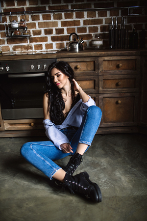Beautiful brunette in the kitchen, modern housewife, fashion, wooden brown furniture, dressed in blue jeans and a blue shirtの写真素材