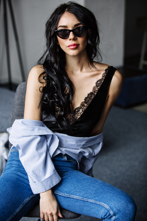 portrait of a beautiful brunette in sunglasses, blue shirt, blue jeans and interior in roomの写真素材