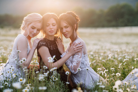 three beautiful girls brunette and blonde, brown-haired woman enjoying Daisy field, nice long dresses, pretty girl relaxing outdoor, having fun, happy young lady and Spring green nature, harmony conceptの写真素材
