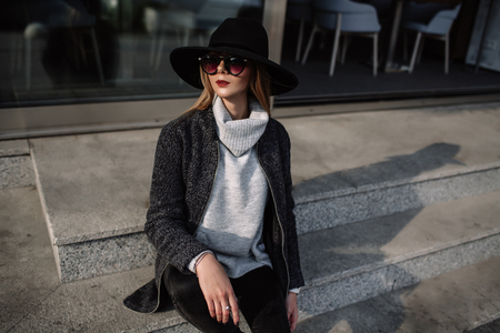 Portrait of a young beautiful fashionable girl wearing sunglasses. Model in a stylish black hat. Harmoniously similar clothes in gray tones. Street style. Women's fashion.の写真素材