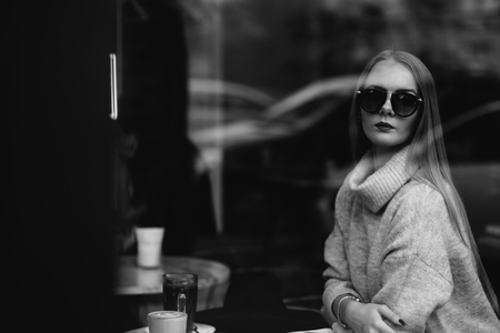 the girl at a table in a cafe drinks coffee, tea, looks out the window, is shot through glass, fashionの写真素材