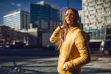 A young European woman, traveling, with long blond hair, wearing a yellow jacket, yellow sunglasses walking down the city center street, street shooting. Even light.の写真素材