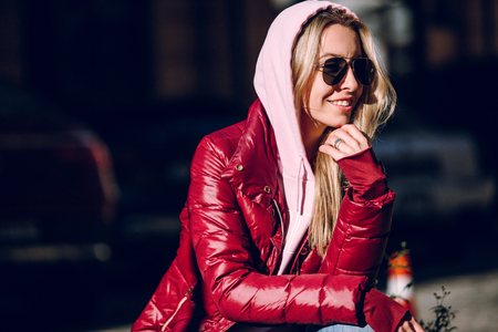 Street fashion concept - portrait of a beautiful girl sitting outside, blue jeans, sunglasses, hard lights, red jacket, autumn weatherの写真素材