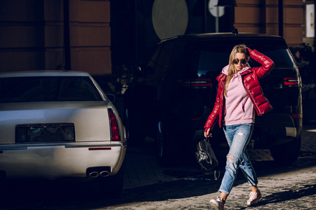 Street fashion concept - portrait of a beautiful girl sitting outside, blue jeans, sunglasses, hard lights, red jacket, autumn weatherの写真素材