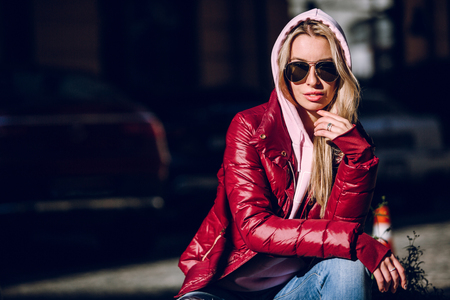 Street fashion concept - portrait of a beautiful girl sitting outside, blue jeans, sunglasses, hard lights, red jacket, autumn weatherの写真素材