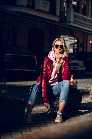 Street fashion concept - portrait of a beautiful girl sitting outside, blue jeans, sunglasses, hard lights, red jacket, autumn weatherの写真素材