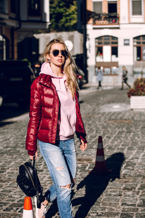 Street fashion concept - portrait of a beautiful girl sitting outside, blue jeans, sunglasses, hard lights, red jacket, autumn weatherの写真素材