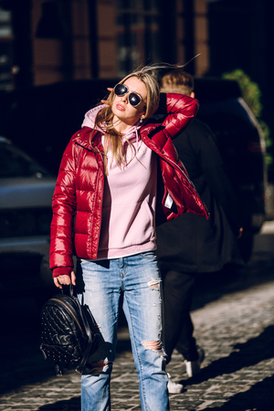 Street fashion concept - portrait of a beautiful girl sitting outside, blue jeans, sunglasses, hard lights, red jacket, autumn weatherの写真素材
