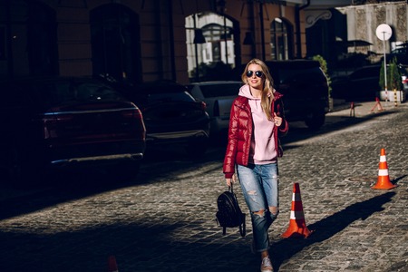 Street fashion concept - portrait of a beautiful girl standing outside, blue jeans, sunglasses, hard lights, red jacket, autumn weatherの写真素材