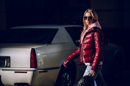 Street fashion concept - portrait of a beautiful girl standing outside, blue jeans, sunglasses, hard lights, red jacket, autumn weatherの写真素材