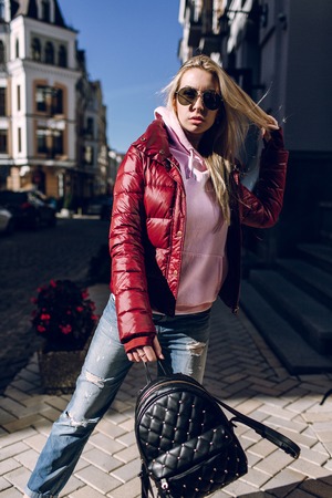 portrait of a beautiful girl standing outside, blue jeans, sunglasses, hard lights, red jacket, autumn weatherの写真素材