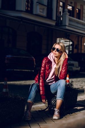 Street fashion concept - portrait of a beautiful girl sitting outside, blue jeans, sunglasses, hard lights, red jacket, autumn weatherの写真素材