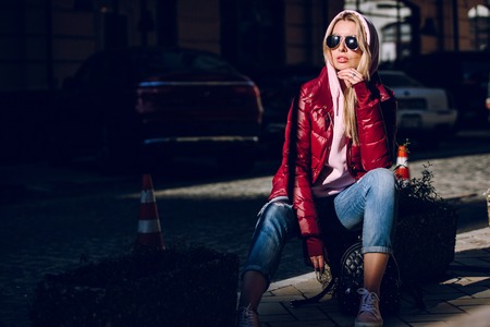 Street fashion concept - portrait of a beautiful girl sitting outside, blue jeans, sunglasses, hard lights, red jacket, autumn weatherの写真素材