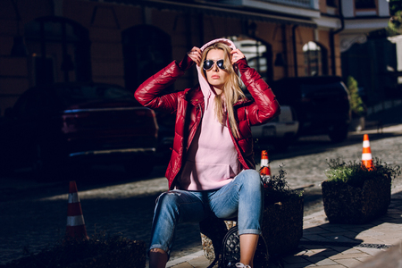 Street fashion concept - portrait of a beautiful girl sitting outside, blue jeans, sunglasses, hard lights, red jacket, autumn weatherの写真素材