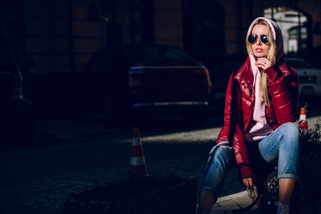 Street fashion concept - portrait of a beautiful girl sitting outside, blue jeans, sunglasses, hard lights, red jacket, autumn weatherの写真素材