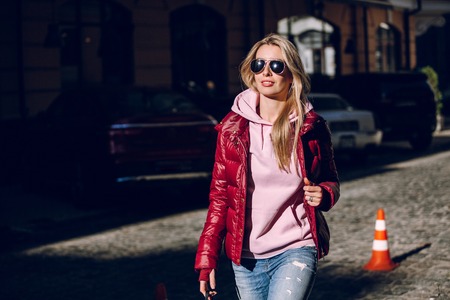 Street fashion concept - portrait of a beautiful girl sitting outside, blue jeans, sunglasses, hard lights, red jacket, autumn weatherの写真素材