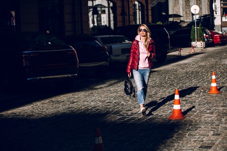 Street fashion concept - portrait of a beautiful girl sitting outside, blue jeans, sunglasses, hard lights, red jacket, autumn weatherの写真素材