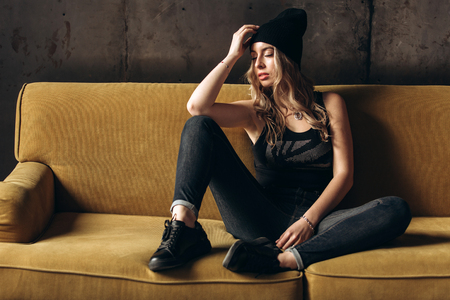 Portrait of a beautiful fashionable woman, blonde, jeans, hat, on a yellow sofaの写真素材