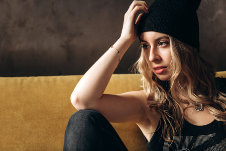 Portrait of a beautiful fashionable woman, blonde, jeans, hat, on a yellow sofaの写真素材