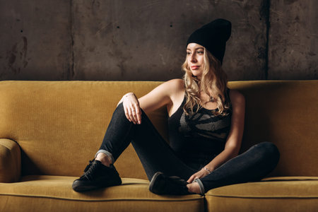 Portrait of a beautiful fashionable woman, blonde, jeans, hat, on a yellow sofaの写真素材