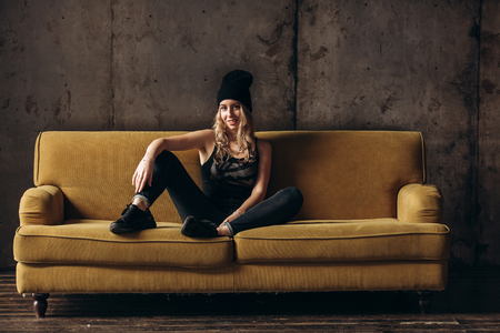 Portrait of a beautiful fashionable woman, blonde, jeans, hat, on a yellow sofaの写真素材