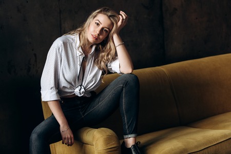 Portrait of a beautiful fashionable woman, blonde, jeans, on a yellow sofaの写真素材