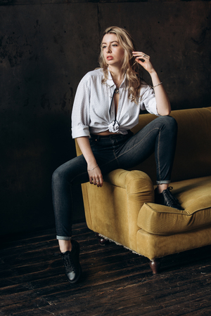 Portrait of a beautiful fashionable woman, blonde, jeans, on a yellow sofaの写真素材