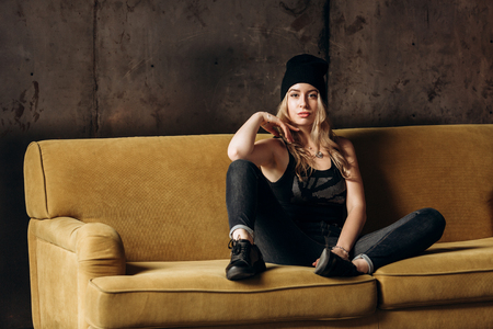 Portrait of a beautiful fashionable woman, blonde, jeans, hat, on a yellow sofaの写真素材