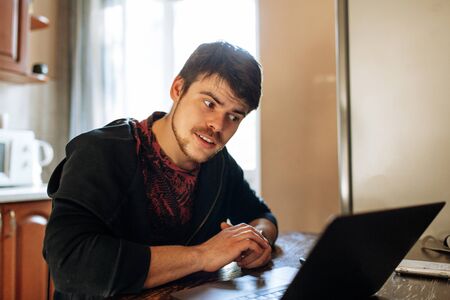 A young bearded hipster man, dressed in a black jacket, sits at home in the kitchen at a table, uses a laptop and talks on his cell phone, calls. Internet marketing, education, e-learning. Startup.の写真素材