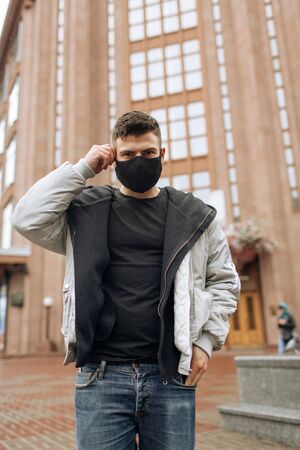 Brunette guy in a black tracksuit puts on a face mask. Black stylish face mask during a pandemic virus crown. Handmade cotton face mask for protection against viruses and bacteria.の写真素材