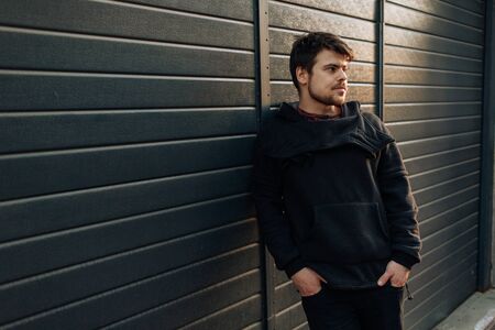Portrait of stylish handsome young man with bristle standing outdoors and leaning on wall. Man wearing black hoodyの写真素材