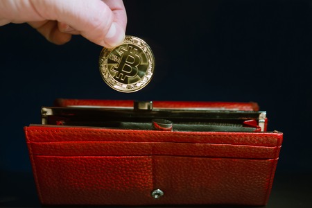 persons hand puts a gold bitcoin in a red wallet that stands on the tableの写真素材