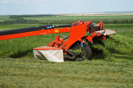 Red tractor trailer cutting grass on the field. The process of mowing clover. Summer work on an...