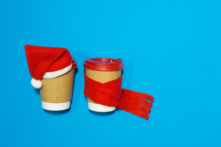 Two paper cups for coffee in a festive outfit from a red cap and a scarf on a blue background....
