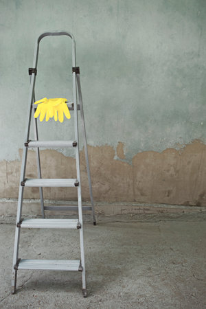 Old folding gray metal ladder and yellow rubber gloves on the background of a dirty construction site. Household repair, construction. Selective focus, artificial noise. Color of the year 2021の写真素材
