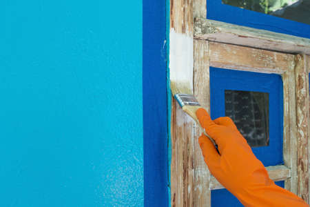 A workers hand in an orange glove paints the window white with a brush. Painting an old wooden frame with sticky masking tape. Household repair, construction. Copy space.の写真素材